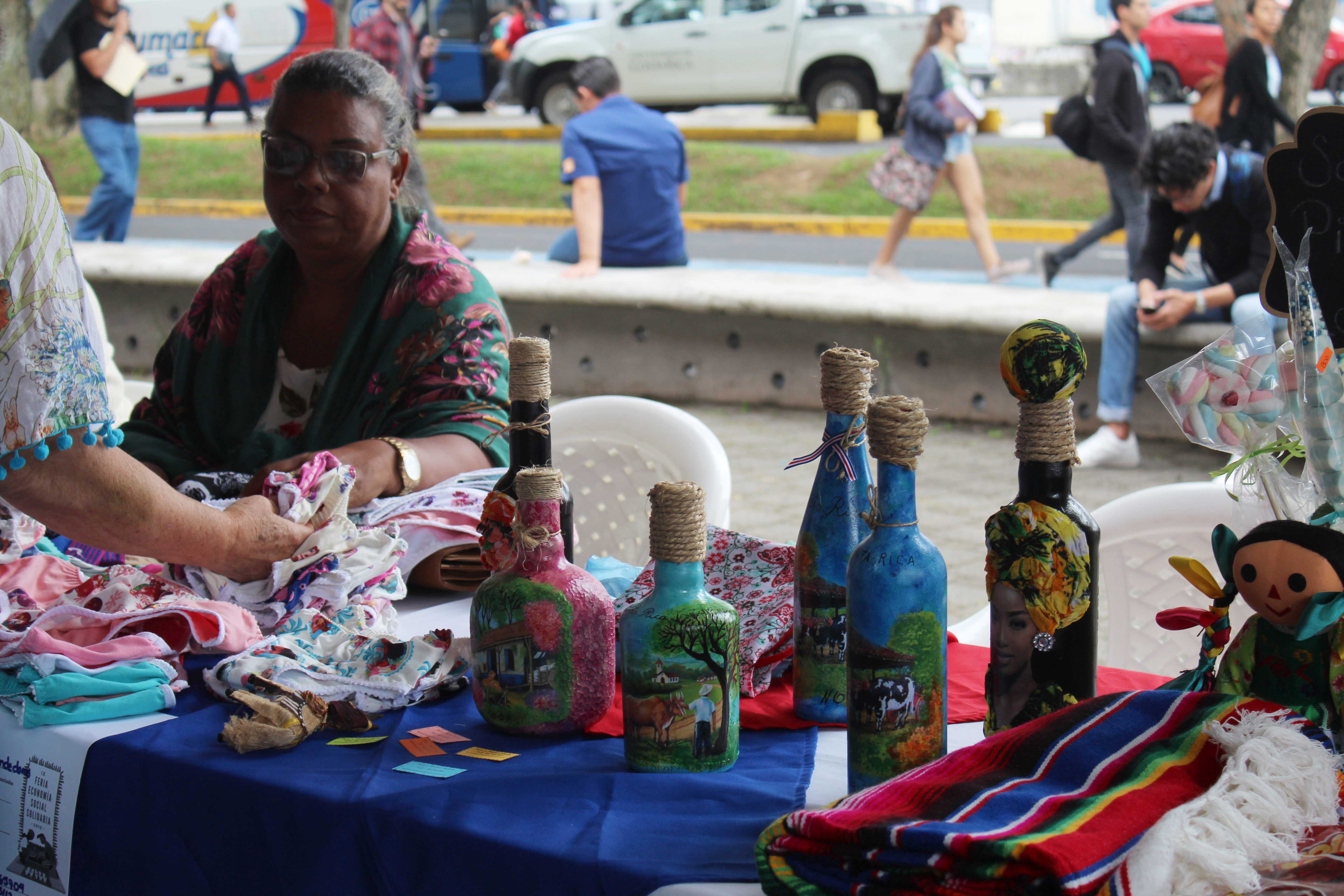 img-galeria-quote-Mujer sentada enseña sus productos a lo largo de la mesa, los cuales incluyen botellas pintadas con distintos paisajes de Costa Rica.