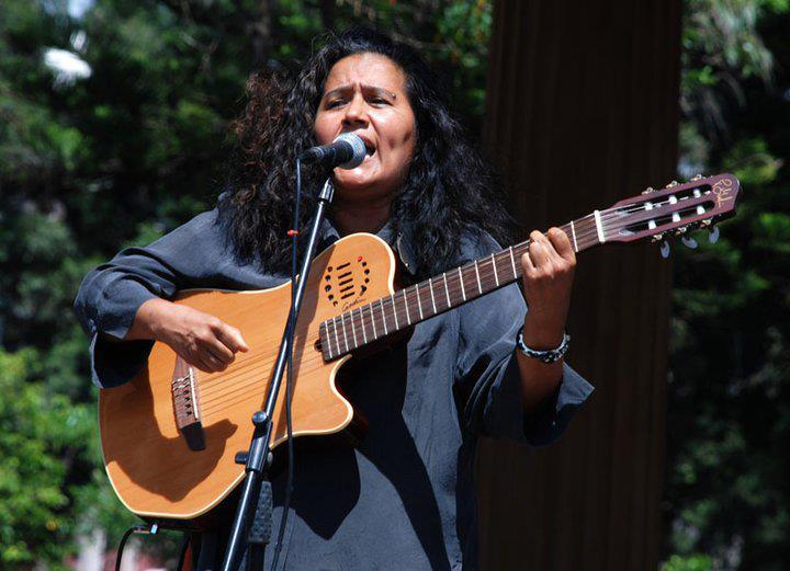Guadalupe Urbina canta frente a un micrófono, mientras toca una guitarra.