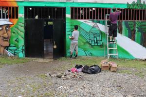 Los artistas Mush y Kein de espaldas trabajando sobre la pared. A la izquierda está la imagen de perfil del líder comunitario “don Roberto”. Fotografía del Programa Kioscos Socio-ambientales. Los artistas Mush y Kein de espaldas trabajando sobre la pared. A la izquierda está la imagen de perfil del líder comunitario “don Roberto”. Fotografía del Programa Kioscos Socio-ambientales.